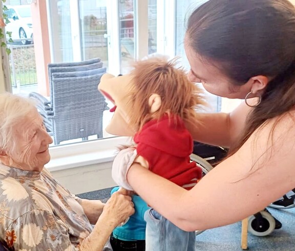 Im Aufenthaltsraum sitzt eine ältere Person auf einem Stuhl und lächelt herzlich. Eine Puppenspielerin steht direkt davor und unterhält sie mit einer Handpuppe – ein Moment voller Freude und Nähe.
