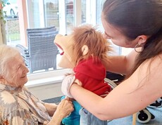 Im Aufenthaltsraum sitzt eine ältere Person auf einem Stuhl und lächelt herzlich. Eine Puppenspielerin steht direkt davor und unterhält sie mit einer Handpuppe – ein Moment voller Freude und Nähe.