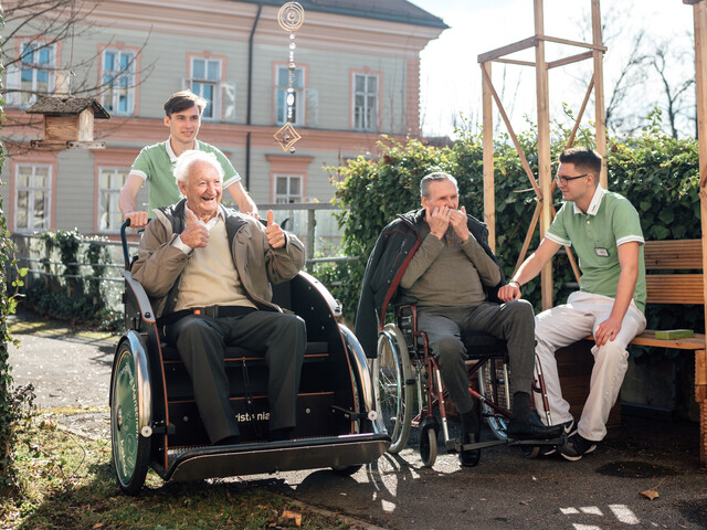 Zwei junge Männer in grünen Poloshirts verbringen Zeit mit zwei älteren Herren im Garten eines Pflegewohnhauses. Links schiebt einer einen lachenden Herrn im Rollstuhl, der beide Daumen hochhält. Rechts sitzt der andere auf einer Holzbank und hört einem Herrn im Rollstuhl zu, der Mundharmonika spielt.