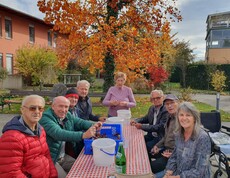 An einem Tisch, im Garten des Pflegewohnhauses, sitzen mehrere Personen – darunter Bewohner sowie freiwillige Mitarbeiterinnen und Mitarbeiter. Gemeinsam bereiten sie Maroni für das Braten vor, indem sie die Schalen sorgfältig einschneiden.