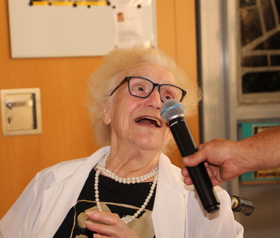 Eine ältere Dame mit Brille und Perlenkette singt in ein Mikrofon, das ihr von einem Herrn gehalten wird.