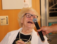 Eine ältere Dame mit Brille und Perlenkette singt in ein Mikrofon, das ihr von einem Herrn gehalten wird.