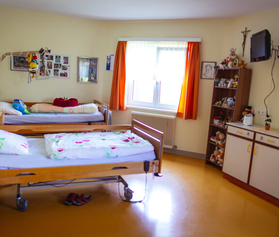 Es befinden sich 2 Betten, ein großes Regal, eine Kommode, ein an der Wand befestigter Fernseher, Bilder, Heizkörper, Fenster mit Vorhängen und 2 Nachtkästchen auf dem Foto.