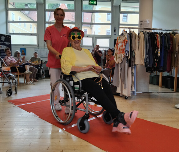 Bewohnerin im Rollstuhl mit Sonnenbrille und gelber Jacke wird über den roten Teppich begleitet.
