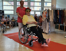 Bewohnerin im Rollstuhl mit Sonnenbrille und gelber Jacke wird über den roten Teppich begleitet.