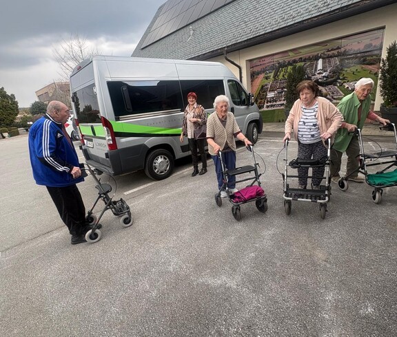 Eine Gruppe von Bewohnerinnen des Pflegewohnhauses ist gemeinsam mit Begleitpersonen auf dem Weg zum Café. Die Gruppe steht vor einem Transporter und bereitet sich mit Rollatoren auf den kurzen Fußweg vor.