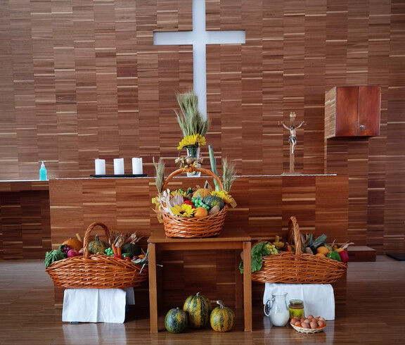  eine Erntedankfest-Dekoration in einer Kirche. Vor einem Holzkreuz sind Körbe und Gaben mit Früchten und Gemüse aufgestellt, die symbolisch für die Ernte und den Dank stehen