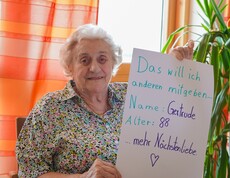Eine Pensionistin hält ein Schild mit ihrem Namen, auf dem steht dass sie anderen mehr Nächstenliebe mitgeben möchte.