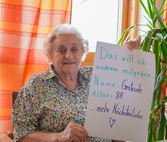 Eine Pensionistin hält ein Schild mit ihrem Namen, auf dem steht dass sie anderen mehr Nächstenliebe mitgeben möchte.