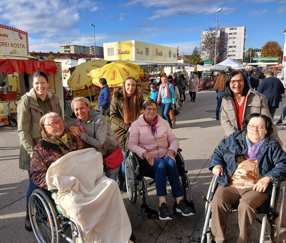 Gruppe von Menschen, einige in Rollstühlen, auf einem Freiluftmarkt mit Ständen und Sonnenschirmen.