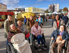Gruppe von Menschen, einige in Rollstühlen, auf einem Freiluftmarkt mit Ständen und Sonnenschirmen.