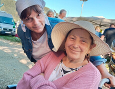 Seniorin im Rollstuhl mit Sonnenhut lächelt, Betreuerin beugt sich freundlich zu ihr