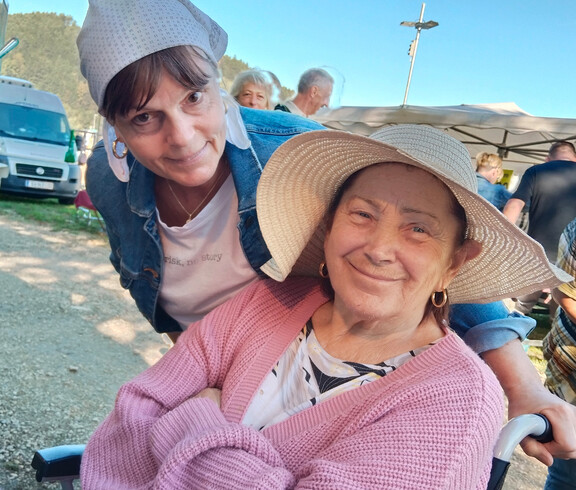 Seniorin im Rollstuhl mit Sonnenhut lächelt, Betreuerin beugt sich freundlich zu ihr