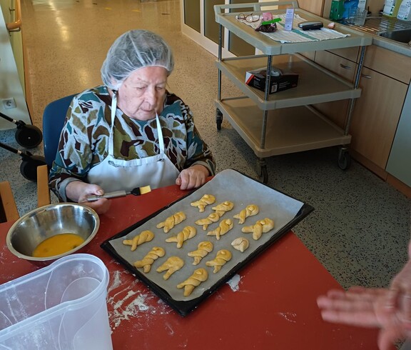 Eine Bewohnerin des Pflegewohnhauses bestreicht frisch geformte Germteig‑Haserl auf einem Backblech mit Eigelb. Vor ihr liegen Backutensilien und Teigreste, während im Hintergrund der gemütliche Küchenbereich zu sehen ist.