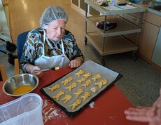 Eine Bewohnerin des Pflegewohnhauses bestreicht frisch geformte Germteig‑Haserl auf einem Backblech mit Eigelb. Vor ihr liegen Backutensilien und Teigreste, während im Hintergrund der gemütliche Küchenbereich zu sehen ist.