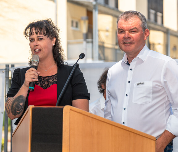 Frau Gaube Claudia und Herr Josef Url stehen am Rednerpult. Frau Gaube hält das Mikrofon und hält eine Begrüßungsrede. Sie trägt ein rot-schwarzes Kleid, er ein weißes Hemd.