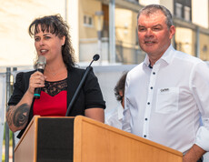 Frau Gaube Claudia und Herr Josef Url stehen am Rednerpult. Frau Gaube hält das Mikrofon und hält eine Begrüßungsrede. Sie trägt ein rot-schwarzes Kleid, er ein weißes Hemd.