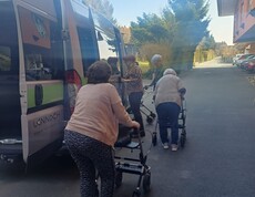 Eine Gruppe von Bewohnerinnen des Pflegewohnhauses ist gemeinsam mit Begleitpersonen auf dem Weg zum Café. Die Gruppe steht vor einem Transporter und bereitet sich mit Rollatoren auf den kurzen Fußweg vor.