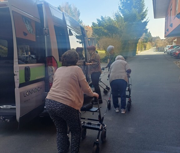 Eine Gruppe von Bewohnerinnen des Pflegewohnhauses ist gemeinsam mit Begleitpersonen auf dem Weg zum Café. Die Gruppe steht vor einem Transporter und bereitet sich mit Rollatoren auf den kurzen Fußweg vor.