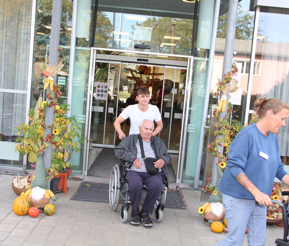 Bewohner*innen werden im Rollstuhl von Mitarbeiter*innen durch den herbstlich mit Kürbissen, Blumen und Dekorationen geschmückten Eingangsbereich des Caritas Pflegewohnhauses ins Freie begleitet, während im Hintergrund weitere Gäste und Bewohner*innen zu sehen sind.