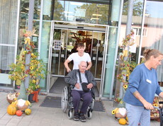 Bewohner*innen werden im Rollstuhl von Mitarbeiter*innen durch den herbstlich mit Kürbissen, Blumen und Dekorationen geschmückten Eingangsbereich des Caritas Pflegewohnhauses ins Freie begleitet, während im Hintergrund weitere Gäste und Bewohner*innen zu sehen sind.