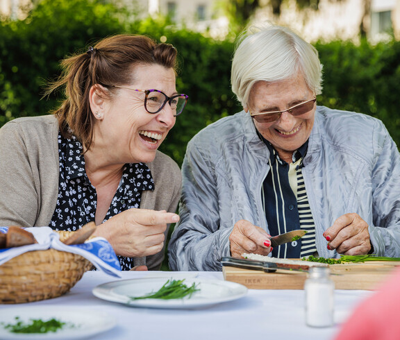 Zwei Damen lachen gemeinsam während sie den Schnittlauch aus dem eigenen Garten klein schneiden.