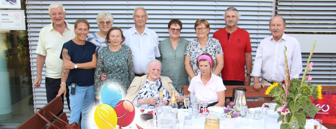 Frau Vacula sitzt an einem großen Tisch, umgeben von ihrer Familie und dem Pflegepersonal – eine herzliche und gemeinschaftliche Atmosphäre.
