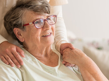 Panoramablick auf eine ältere Dame im Rollstuhl. Hinter ihr steht ein unterstützender Freiwilliger, der ihre linke Hand hält.