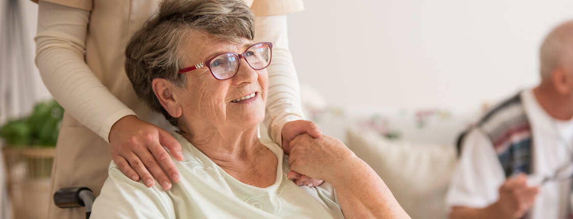 Panoramablick auf eine ältere Dame im Rollstuhl. Hinter ihr steht ein unterstützender Freiwilliger, der ihre linke Hand hält.