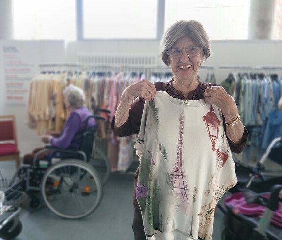 Bewohnerin hält ein helles Oberteil mit Paris‑Motiv vor Kleiderständern