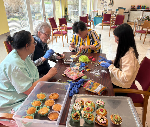 Bewohner*innen gestalten gemeinsam Osterdekoration und Süßspeisen an einem Tisch im Gemeinschaftsraum.