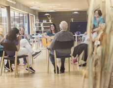 Das Bild zeigt eine Gruppenaktivität im Tageszentrum, bei der mehrere ältere Menschen im Kreis zusammensitzen und gemeinsam musizieren und sich austauschen, begleitet von Betreuungspersonen in einem hellen, offenen Raum.