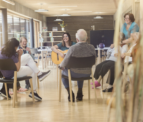 Das Bild zeigt eine Gruppenaktivität im Tageszentrum, bei der mehrere ältere Menschen im Kreis zusammensitzen und gemeinsam musizieren und sich austauschen, begleitet von Betreuungspersonen in einem hellen, offenen Raum.