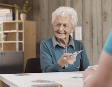 Das Bild zeigt eine ältere Person, die an einem Tisch im Tageszentrum sitzt und einer gegenüberstehenden Person Spielkarten reicht, wobei der helle Innenraum mit Regalen und Holzelementen eine ruhige, gemeinschaftliche Umgebung für betreute Aktivitäten bildet.