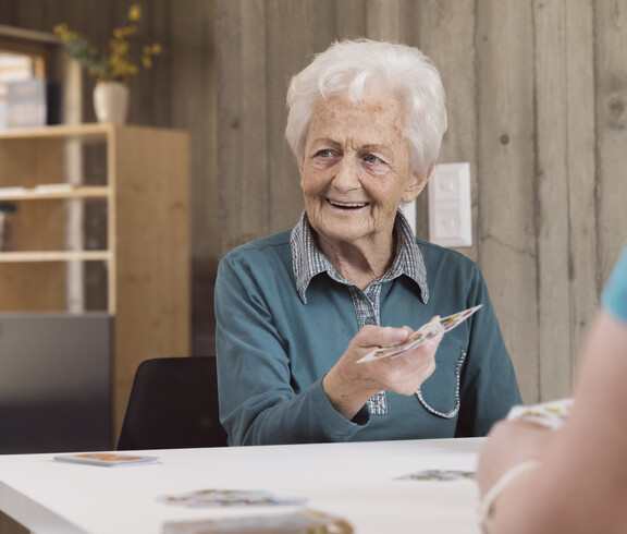 Das Bild zeigt eine ältere Person, die an einem Tisch im Tageszentrum sitzt und einer gegenüberstehenden Person Spielkarten reicht, wobei der helle Innenraum mit Regalen und Holzelementen eine ruhige, gemeinschaftliche Umgebung für betreute Aktivitäten bildet.