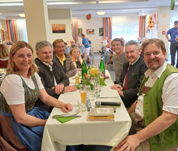 Im Gemeinschaftsraum sitzen die Gäste an einem gedeckten Tisch. Unter ihnen befinden sich Bürgermeister Josef Niggas, Caritaspräsidentin und -direktorin Nora Tödtling-Musenbichler sowie Max Tödtling, Moderator der Feier und ehemaliger Hausleiter. Im Hintergrund sind weitere Tische und der Raum sichtbar.