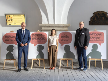Eine Frau in der Mitte, ein Mann rechts, ein Mann links von ihr stehend vor einer Reihe von Schildern mit Sprechblasen.