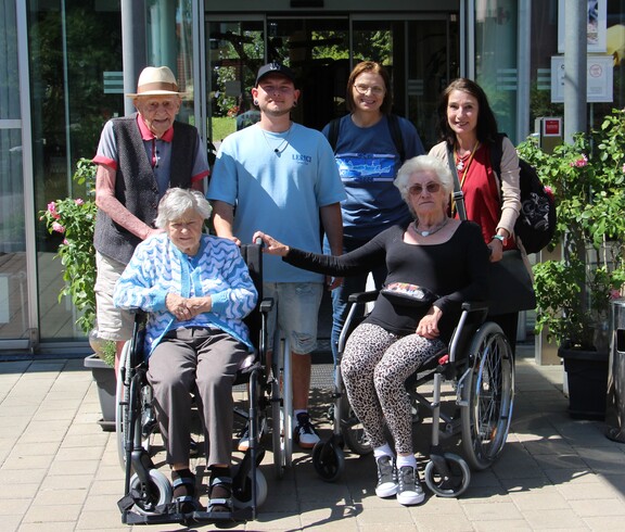 Zwei Bewohnerinnen sitzen nebeneinander in Rollstühlen. Hinter ihnen steht ein weiterer Bewohner, der sich leicht an einem der Rollstühle festhält. Direkt daneben befindet sich Pflegepersonal, das aufmerksam und unterstützend wirkt. Die Gruppe steht vor dem Eingang eines Museums.