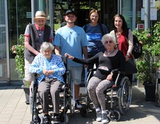 Zwei Bewohnerinnen sitzen nebeneinander in Rollstühlen. Hinter ihnen steht ein weiterer Bewohner, der sich leicht an einem der Rollstühle festhält. Direkt daneben befindet sich Pflegepersonal, das aufmerksam und unterstützend wirkt. Die Gruppe steht vor dem Eingang eines Museums.
