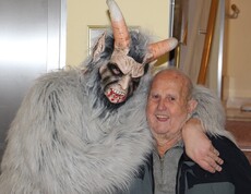 Krampus mit hellgrauem Fell steht neben einem Bewohner des Pflegewohnhauses und legt freundlich seine Hand über dessen Schulter.