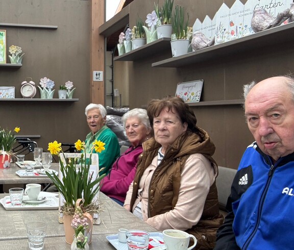 Bewohnerinnen und Bewohner des Pflegewohnhauses sitzen gemeinsam mit Mitgliedern der Frauenbewegung in einem Gartencafé an einem gedeckten Tisch. Vor ihnen stehen Kaffeetassen, Blumenarrangements und kleine Dekorationen. Die Gruppe genießt in gemütlicher Atmosphäre einen gemeinsamen Ausflug.