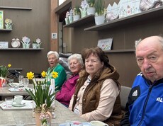 Bewohnerinnen und Bewohner des Pflegewohnhauses sitzen gemeinsam mit Mitgliedern der Frauenbewegung in einem Gartencafé an einem gedeckten Tisch. Vor ihnen stehen Kaffeetassen, Blumenarrangements und kleine Dekorationen. Die Gruppe genießt in gemütlicher Atmosphäre einen gemeinsamen Ausflug.