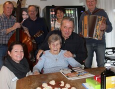 Eine Bewohnerin feiert im Pflegewohnhaus ihren 102. Geburtstag. Vor ihr steht eine Torte mit der Zahl 102. Neben ihr befindet sich die Hausleiterin; im Hintergrund sind Familienangehörige und Musikanten zu sehen.