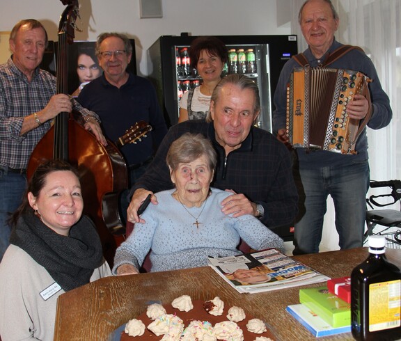 Eine Bewohnerin feiert im Pflegewohnhaus ihren 102. Geburtstag. Vor ihr steht eine Torte mit der Zahl 102. Neben ihr befindet sich die Hausleiterin; im Hintergrund sind Familienangehörige und Musikanten zu sehen.