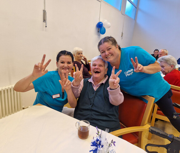 Zwei Pflegekräfte und eine Seniorin posieren lachend mit Friedenszeichen