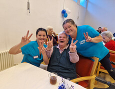 Zwei Pflegekräfte und eine Seniorin posieren lachend mit Friedenszeichen