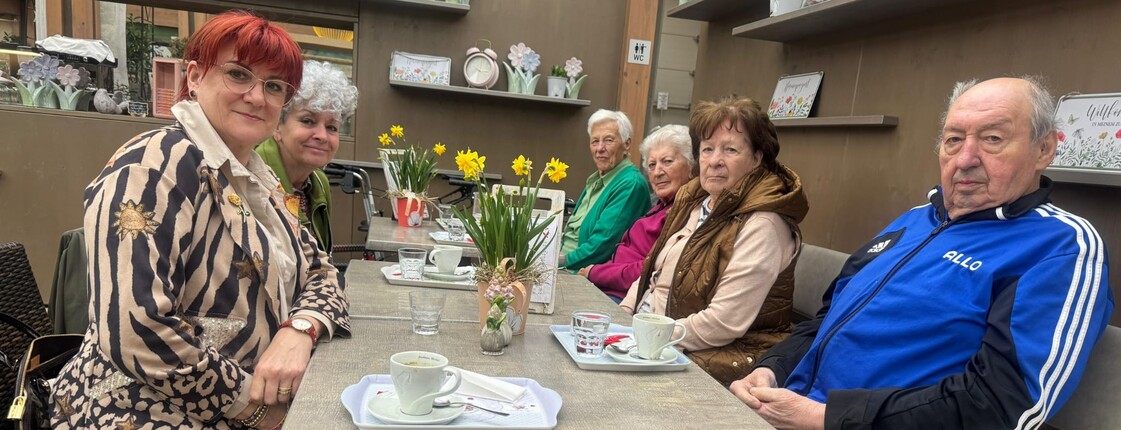 Bewohnerinnen und Bewohner des Pflegewohnhauses sitzen gemeinsam mit Mitgliedern der Frauenbewegung in einem Gartencafé an einem gedeckten Tisch. Vor ihnen stehen Kaffeetassen, Blumenarrangements und kleine Dekorationen. Die Gruppe genießt in gemütlicher Atmosphäre einen gemeinsamen Ausflug.