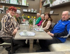 Bewohnerinnen und Bewohner des Pflegewohnhauses sitzen gemeinsam mit Mitgliedern der Frauenbewegung in einem Gartencafé an einem gedeckten Tisch. Vor ihnen stehen Kaffeetassen, Blumenarrangements und kleine Dekorationen. Die Gruppe genießt in gemütlicher Atmosphäre einen gemeinsamen Ausflug.