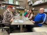 Bewohnerinnen und Bewohner des Pflegewohnhauses sitzen gemeinsam mit Mitgliedern der Frauenbewegung in einem Gartencafé an einem gedeckten Tisch. Vor ihnen stehen Kaffeetassen, Blumenarrangements und kleine Dekorationen. Die Gruppe genießt in gemütlicher Atmosphäre einen gemeinsamen Ausflug.