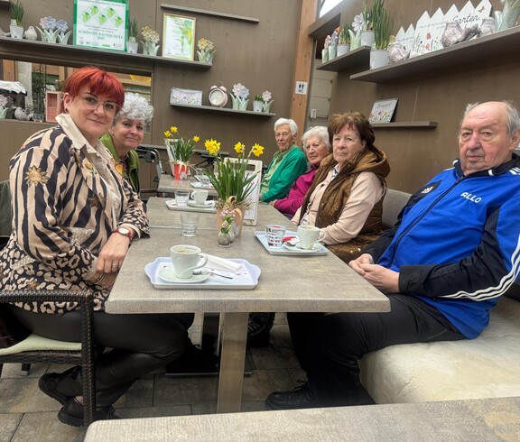 Bewohnerinnen und Bewohner des Pflegewohnhauses sitzen gemeinsam mit Mitgliedern der Frauenbewegung in einem Gartencafé an einem gedeckten Tisch. Vor ihnen stehen Kaffeetassen, Blumenarrangements und kleine Dekorationen. Die Gruppe genießt in gemütlicher Atmosphäre einen gemeinsamen Ausflug.